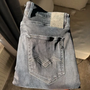 Svarta Replay jeansbyxor - Säljer ett par svarta jeans från Replay med klassisk femficksdesign och snygga detaljer på bakfickorna. Byxorna har normal passform och är tillverkade i stretchigt denim för extra komfort. Perfekta för dig som gillar stilrena och tidlösa jeans.