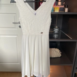 Vit spetsklänning med v-ringning - Säljer en söt vit klänning med v-ringning och spets upptill. Kjolen är slät och faller fint, och klänningen är ärmlös. Perfekt för sommarens tillfällen eller studentfirande.