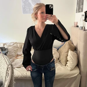 Svart omlott bodysuit med knappar - Säljer en svart bodysuit med omlott-skärning och långa ärmar. Ärmarna har dekorativa silverfärgade knappar längs manschetterna som ger en cool detalj. Perfekt att styla med jeans eller kjol. Den är i st M men passar mig som är XS/S