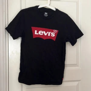 Svart t-shirt från Levi's - Svart t-shirt från Levi's med klassisk röd och vit logga framtill. Enkel och stilren design med rund hals och korta ärmar. Perfekt till jeans eller shorts. STORLEK XS men stor i storlek 