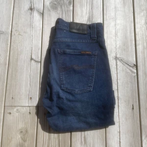 Nudie jeans - Säljer ett par mörkblå jeansbyxor från Nudie  med klassisk femficksdesign och raka ben. Byxorna har en diskret nudie-logga på bakfickan och är tillverkade i ett kvalitets denimtyg för extra komfort. Model Lean Dean! Midja ca 37cm