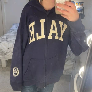 Mörkblå hoodie med YALE-tryck - Säljer en mörkblå zip hoodie med dragkedja och stor vit YALE-text på bröstet. Tröjan har huva, två fickor fram och ribbade muddar. Perfekt för en avslappnad och cool stil.