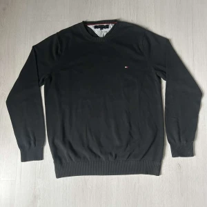 Svart tröja från Tommy Hilfiger - Klassisk svart tröja från Tommy Hilfiger med rund halsringning och diskret logga på bröstet. Tröjan har långa ärmar och ribbade muddar vid ärmslut och nederkant. Perfekt för en stilren och enkel look. 