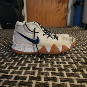 Kyrie 4 uncle drew - Säljer ett par vita Nike sneakers med blå swoosh och unika bruna sula med orange detaljer. Skorna har snörning och högt skaft, perfekta för basket eller streetstyle. Mönstrad yttersula och stänkmönster på mellansulan ger extra attityd.