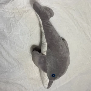 Delfin gosedjur  - Det här är ett gosedjur som föreställer en delfin, inte en bok. Den har en mjuk grå ovansida och vit undersida, med blå ögon. Perfekt för dig som gillar havsdjur och vill ha ett mysigt sällskap.