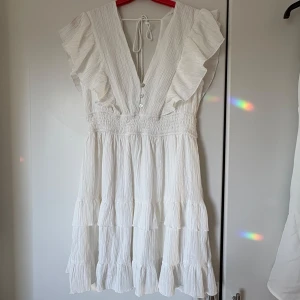 Vit volangklänning med knyt i ryggen - Superfin vit klänning med volanger och djup v-ringning framtill. Klänningen har knappar framtill, smock i midjan och knytband i ryggen. Perfekt för sommare och studenttiderna⭐️🌸