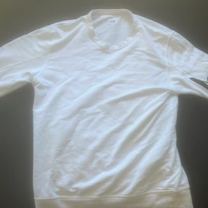 Vit jumper från cp company - Säljer en vit jumper från C.P. Company med klassisk passform. Tröjan har rund halsringning och en ikonisk linsdetalj på ärmen. Perfekt för en clean och stilren look.