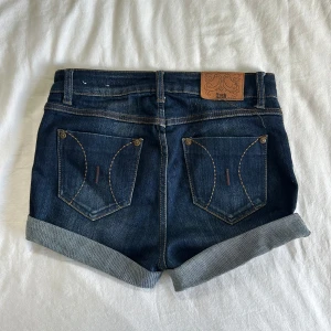 Mörkblå jeansshorts från Flash Jeans - Snygga mörkblå jeansshorts från Flash Jeans med uppvikta benslut och klassiska bakfickor med dekorativa sömmar. Modellen heter Kelly och shortsen har normal passform och knappgylf. Perfekta för sommaren!