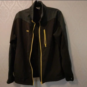Svart jacka med gula detaljer från Didriksons - Säljer en svart jacka från Didriksons med gula dragkedjor och detaljer. Jackan har hög krage, lång ärm och en bröstficka med dragkedja. Perfekt för utomhusaktiviteter och har en sportig look.