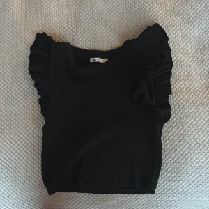 Svart croppad topp med volangärmar från Zara - Säljer en svart croppad topp från Zara med volangdetaljer på axlarna. Toppen har rund halsringning och är tillverkad i ett mjukt material. Perfekt för att lyfta en enkel outfit.