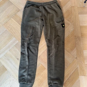 Olivgröna mjukisbyxor från Lyle & Scott Junior - Säljer ett par olivgröna mjukisbyxor från Lyle & Scott Junior med resår i midjan och muddar vid bensluten. Byxorna har en ficka på benet med Lyle & Scott-logga i gult och svart. Perfekta för en avslappnad stil.