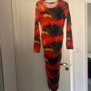 Färgstark långklänning med drapering - Långklänning med färgstarkt mönster i rött, orange, grönt och svart. Klänningen har skrynkligeffekt längs sidorna. 