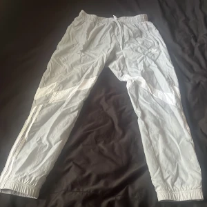 Ljusgrå träningsbyxor från Adidas - Säljer ett par ljusgrå träningsbyxor (tunna byxor) från Adidas med klassiska vita ränder längs sidorna och dragsko i midjan. Byxorna har resår vid bensluten och dragkedjefickor. Perfekta för sport eller chill. Samt dragkedja vid benen.