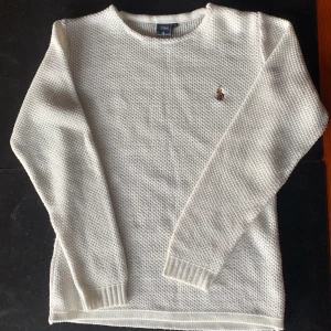 Vit stickad tröja från Polo Ralph Lauren - Klassisk vit stickad tröja från Polo Ralph Lauren med rund halsringning och broderad logga på bröstet. Tröjan har lång ärm och ribbade muddar vid ärmslut och nederkant. Perfekt för lager-på-lager under kyliga dagar. 