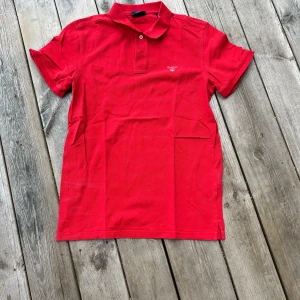 Röd pikétröja från Gant - Säljer en klassisk röd pikétröja från Gant med krage och knappslå. Tröjan har korta ärmar och liten broderad logga på bröstet. Perfekt för en stilren och avslappnad look.