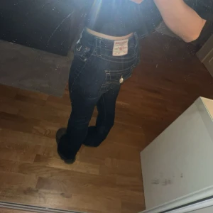 Blå bootcut jeans från True Religion - Säljer ett par klassiska blå jeans från True Religion med bootcut passform. Jeansen har dekorativa sömmar och fickor med lock bak samt tydlig logga på bakfickan. 