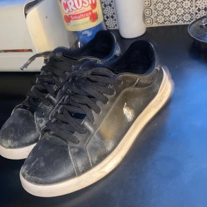 Ralph Lauren skor  - Svarta sneakers från Polo Ralph Lauren med vit logga på sidan. Skorna har snörning, rund tå och vit platt sula. Ovandelen är i skinn och insidan i textil, vilket gör dem både snygga och bekväma.