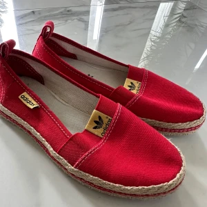 Röda espadrillos från Adidas - Säljer ett par snygga röda espadrillos från Adidas med klassisk flätad sula och rund tå. Skorna har tygöverdel och små gula Adidas-loggor på sidan och framtill. Perfekta för varma dagar och enkel att matcha till sommarens outfits.