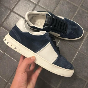 Valentino Flycrews  - Säljer ett par snygga sneakers från Valentino i blå mocka med vita detaljer och svarta skosnören. Skorna har platt sula och små nitar på hälen för en extra touch. Perfekta för dig som gillar stilrena men unika sneakers.