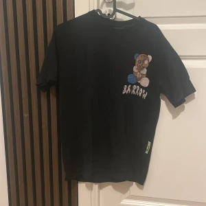 Svart t-shirt med nalletryck från Barrow - Cool svart t-shirt från Barrow med ett stort tryck av en nalle på både fram- och baksidan. Framsidan har även Barrow-loggan och baksidan har text och ett smiley-tryck. Perfekt för dig som gillar streetwear och unika prints.