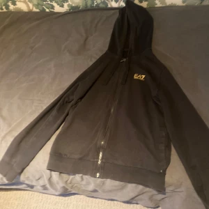 Svart zip hoodie från EA7 Emporio Armani - Svart hoodie med dragkedja från EA7 Emporio Armani. Tröjan har huva, ribbade muddar och en liten gul logga på bröstet samt dekorativt märke baktill. Perfekt för en sportig och avslappnad stil. Tröjan är i storleken S och jag säljer den eftersom den inte passar mig längre.