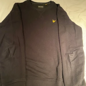 Svart sweatshirt från Lyle & Scott - Säljer en svart sweatshirt från Lyle & Scott med klassisk rund halsringning och gul broderad logga på bröstet. Tröjan har långa ärmar och ribbade muddar vid ärmslut och nederkant. Perfekt för en stilren och enkel look.