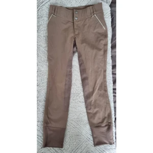 Beige ridbyxor från CRW med dragkedjedetaljer - Snygga beige ridbyxor från CRW med förstärkning på insidan av benen och dragkedjor vid fickorna. Byxorna har två knappar framtill och bälteshällor. Stl 160