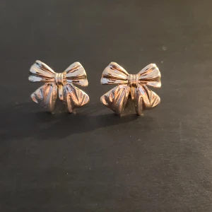 Guldiga örhängen i form av rosetter, clipons - Säljer ett par söta örhängen i guldmetall formade som rosetter. Perfekta för att ge en gullig touch till din outfit. Små och nätta, passar till många olika stilar.