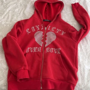 Röd throtherz zip hoodie - Röd hoodie med dragkedja framtill och vit text samt hjärtmotiv på bröstet. Trycket säger 'Loyalty Over Love' och har ett 