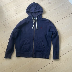 Mörkblå hoodie med dragkedja från Polo Ralph Lauren - Säljer en mörkblå hoodie från Polo Ralph Lauren i storlek L. Tröjan har dragkedja, huva med vita snören och en röd broderad logga på bröstet. Skick 8/10.