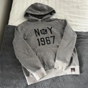 Grå hoodie från Polo Ralph Lauren - Säljer en grå hoodie från Polo Ralph Lauren med svart tryck 'N RLY 1967' på bröstet. Tröjan har huva, magficka och ribbade muddar vid ärmslut och nederkant. Perfekt för en avslappnad stil.