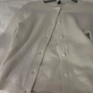 Vit kofta från H&M - Säljer en klassisk vit kofta från H&M med rund halsringning och knäppning framtill. Koftan är stickad i mjukt material och har långa ärmar samt ribbade muddar vid ärmslut och nederkant. Perfekt basplagg till garderoben.