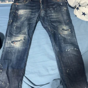 Blå distressed jeans från Dsquared2 storlek 44 men passar  som storlek S  - Säljer ett par blå jeans från Dsquared2 med slitna detaljer, hål och färgstänk. Jeansen har klassisk femficksdesign och läderpatch med logga bak. Raka ben och normal passform. Perfekta för dig som gillar en edgy streetstil.