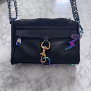 Svart axelväska med regnbågsdetaljer från Rebecca Minkoff - Cool svart axelväska i skinn från Rebecca Minkoff med regnbågsfärgade metalldetaljer, kedjerem och blixtformad berlock. Väskan har dragkedja upptill, dekorativ krok framtill och små nitar på undersidan. Perfekt för att lyfta vilken outfit som helst.