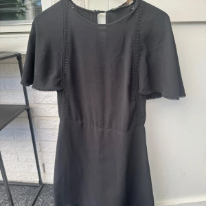 Svart kortklänning med volangärmar - Säljer en svart kortklänning med fina volangärmar och rund halsringning. Klänningen har dekorativa spetsdetaljer längs axlarna och är tillverkad i ett mjukt material. Perfekt för dig som gillar en stilren look med en twist.