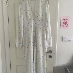 Vit långklänning med blå blommor från Adoore - Vit långklänning från Adoore med små blå blommor, v-ringning och knappar framtill. Klänningen är långärmad. Perfekt för somriga tillfällen. Använd endast till midsommar. 