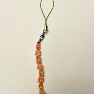 Mobilsmycke blommor  mobilband med pärlor - Sött mobilband med små pärlor i form av rosa och orange blommor med gröna detaljer. Bandet har en grön snodd och metallfäste för enkel fastsättning på mobilen eller väskan. Handgjord. Ca 12 cm totalt. Delen med blommorna ca 7 cm.