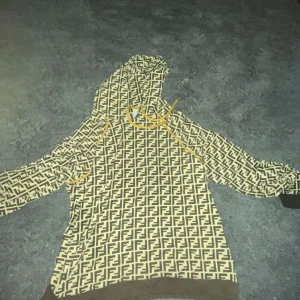 Beige och brun hoodie med Fendi-mönster - Säljer en beige och brun hoodie från Fendi med det klassiska FF-mönstret över hela tröjan. Tröjan har huva med gula snören och muddar i brunt vid ärmslut och nederkant. Perfekt statement-plagg för dig som gillar lyxiga märken.