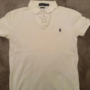 Vit pikétröja från Polo Ralph Lauren - Vit pikétröja från Polo Ralph Lauren med klassisk krage och knappar framtill. Tröjan har korta ärmar och den ikoniska broderade loggan i mörkblått på bröstet. Modellen är custom slim fit. 