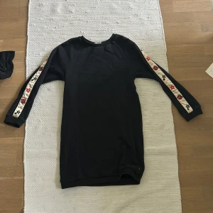 Svart klänning med mönstrade ärmar från Scotch & Soda - Svart klänning från Scotch & Soda med långa ärmar. Ärmarna har en beige rand med broderade stjärnor och rosor i rött och svart. Mjuk och bekväm modell, perfekt för en cool och avslappnad stil.