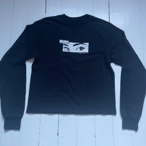 Svart sweatshirt med ögonmotiv - Säljer en svart sweatshirt med tryck av ögon och text på bröstet. Tröjan har rund halsringning och långa ärmar. Perfekt för dig som gillar grafiska detaljer och en stilren look.