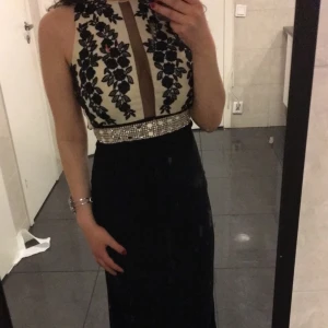 Långklänning - Säljer en elegant svart långklänning med ärmlös design. Överdelen är vit med svarta blommiga broderier och en transparent v-ringad detalj framtill. Midjan pryds av ett brett band med pärlor och glittrande stenar. Perfekt för bal, bröllop eller annan fest.