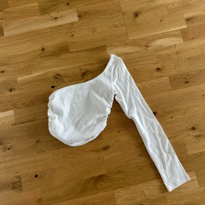 Vit oneshoulder topp från Zara - Säljer en vit ribbad oneshoulder topp från Zara. Toppen har en lång ärm och är tillverkad i ett stretchigt ribbat material som sitter snyggt på kroppen.