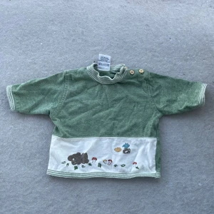 Grön bomullströja med broderi och knappar - Söt ljusgrön tröja i 100% bomull med vita ränder på krage och ärmslut. Tröjan har tre träfärgade knappar på axeln och ett vitt band nertill med broderade djur, svampar och blad. Perfekt för en mysig och lekfull stil.