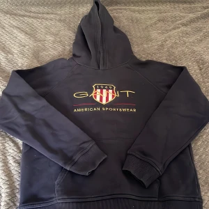 Mörkblå hoodie från Gant - Säljer en mörkblå hoodie från Gant med broderad logga och sköld på bröstet. Tröjan har huva och magficka, samt ribbade muddar vid ärmslut och nederkant. Perfekt för en sportig och avslappnad stil.
