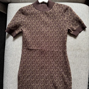 Brun kortärmad klänning från Michael Kors - Säljer en brun kortärmad klänning från Michael Kors med MK-logga som mönster över hela plagget. Klänningen har ribbad hög krage och ärmar, och en figurnära passform. Perfekt för dig som gillar stilrena och trendiga plagg.
