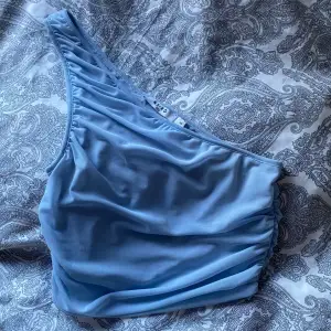 Fin ljusblå oneshoulder topp från NA-KD. Perfekt för en stilren look med sin asymmetriska design och mjuka material. Toppen har en rynkad detalj på sidan som ger en extra touch. OBS! Den har en sminkfläck, syns inte så mycket när man har på sig den, syns på bild 3