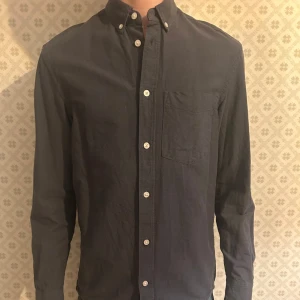 Svart långärmad skjorta med knappar - Snygg svart långärmad skjorta med klassisk krage och knappar hela vägen ner. Skjortan har en bröstficka och är perfekt för en stilren look. Storlek XS. Nypris cirka 300kr. Hör av dig vid fundering mvh Levi!