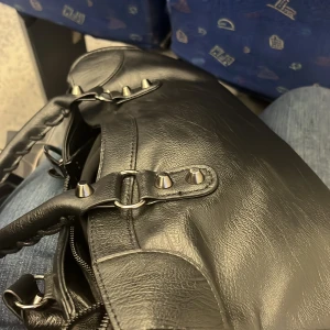 Svart axelväska med nitar - Snygg svart axelväska i skinn med coola nitar och flätade detaljer. Väskan har en dragkedja och flera fack för praktisk förvaring. Perfekt för att ge din outfit en edgy touch.