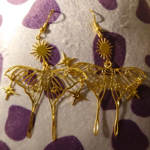 Guldiga mal örhängen med stjärndetaljer - Unika guldiga örhängen i form av malar med sol- och stjärndetaljer. Perfekta för att ge en magisk touch till din stil. Lätta och eleganta, dessa örhängen är ett måste för smyckesälskare.